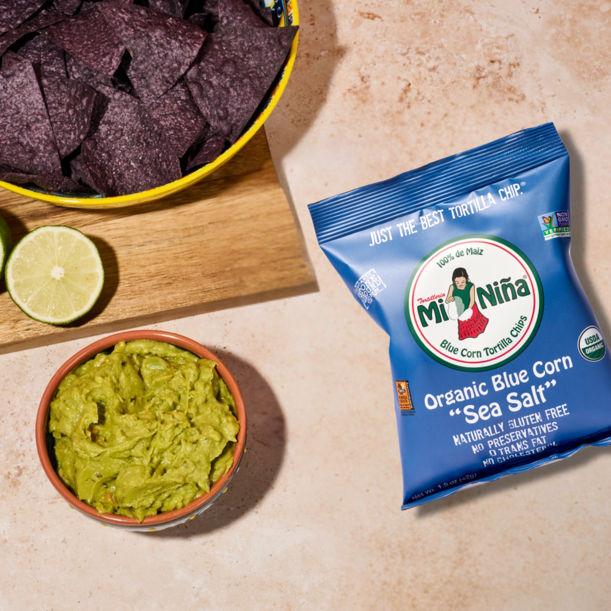 A bowl of crunchy Mi Niña Tortilla Organic Blue Corn “Sea Salt” Tortilla Chips (from the 24 Pack / 1.5 oz), served with sliced lime and creamy guacamole in a dish, alongside a bag of chips displayed on a beige surface.