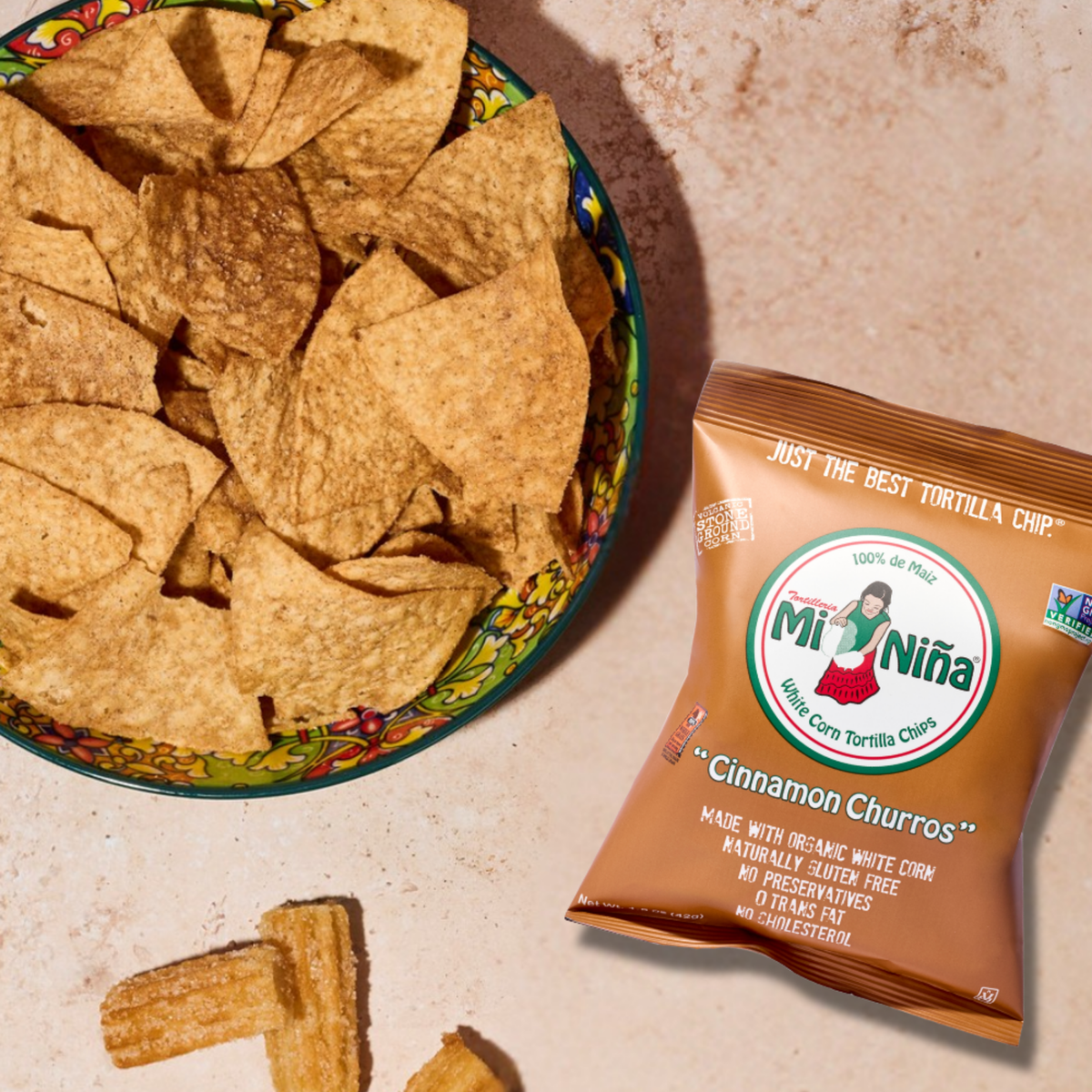 A vibrant bowl brimming with tortilla chips sits beside a bag of Mi Niña Tortilla Cinnamon “Churros” Tortilla Chips (24 Pack / 1.5 oz) and two loose churro chips on a light surface, showcasing why these treats from Mi Niña Tortilla are such a delicious choice.