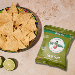 A bowl of tortilla chips, offering the crisp flavor of white corn and a hint of olive oil, sits next to a green bag of Mi Niña Tortilla Sea Salt Made with Olive Oil chips (24 Pack / 1.5 oz) and two lime halves on a textured surface.