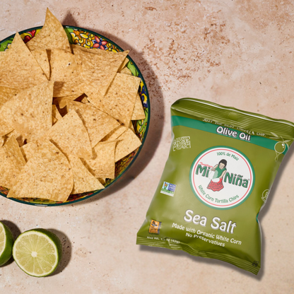 A bowl of tortilla chips, offering the crisp flavor of white corn and a hint of olive oil, sits next to a green bag of Mi Niña Tortilla Sea Salt Made with Olive Oil chips (24 Pack / 1.5 oz) and two lime halves on a textured surface.