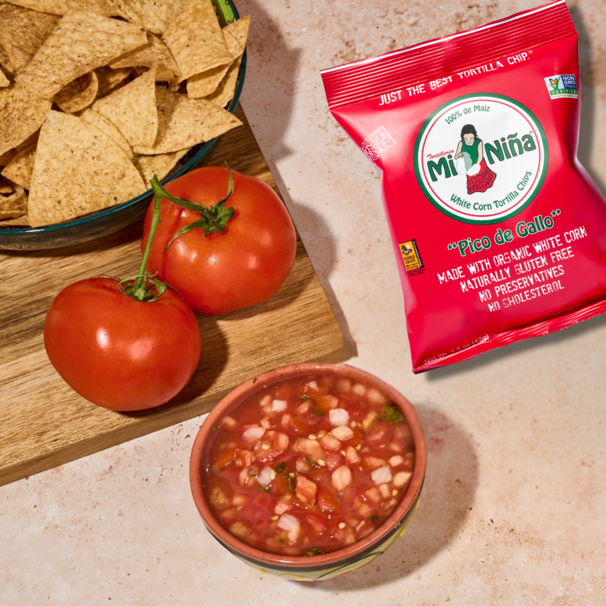 A 1.5 oz bag of Mi Niña Tortilla Pico de Gallo Tortilla Chip Snack Bags next to a bowl of pico de gallo, two whole tomatoes, and a bowl of white corn tortilla chips.