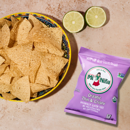 A bowl of thin & crispy tortilla chips sits next to two lime halves and a bag of Mi Niña Tortilla Organic Thin & Crispy Sea Salt – 24 Pack / 1.5 oz chips on a beige surface.