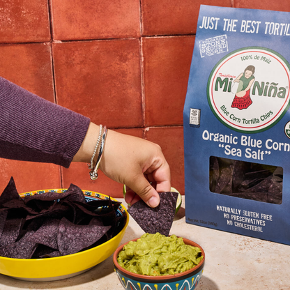A hand dips a crunchy Mi Niña Tortilla Organic Blue Corn “Sea Salt” Tortilla Chip into creamy guacamole. Nearby sit a bowl of chips and a bag labeled “Organic Blue Corn ‘Sea Salt’ Tortilla Chip - 9 Pack / 12 oz” from Mi Niña Tortilla, all set against a red tiled background.