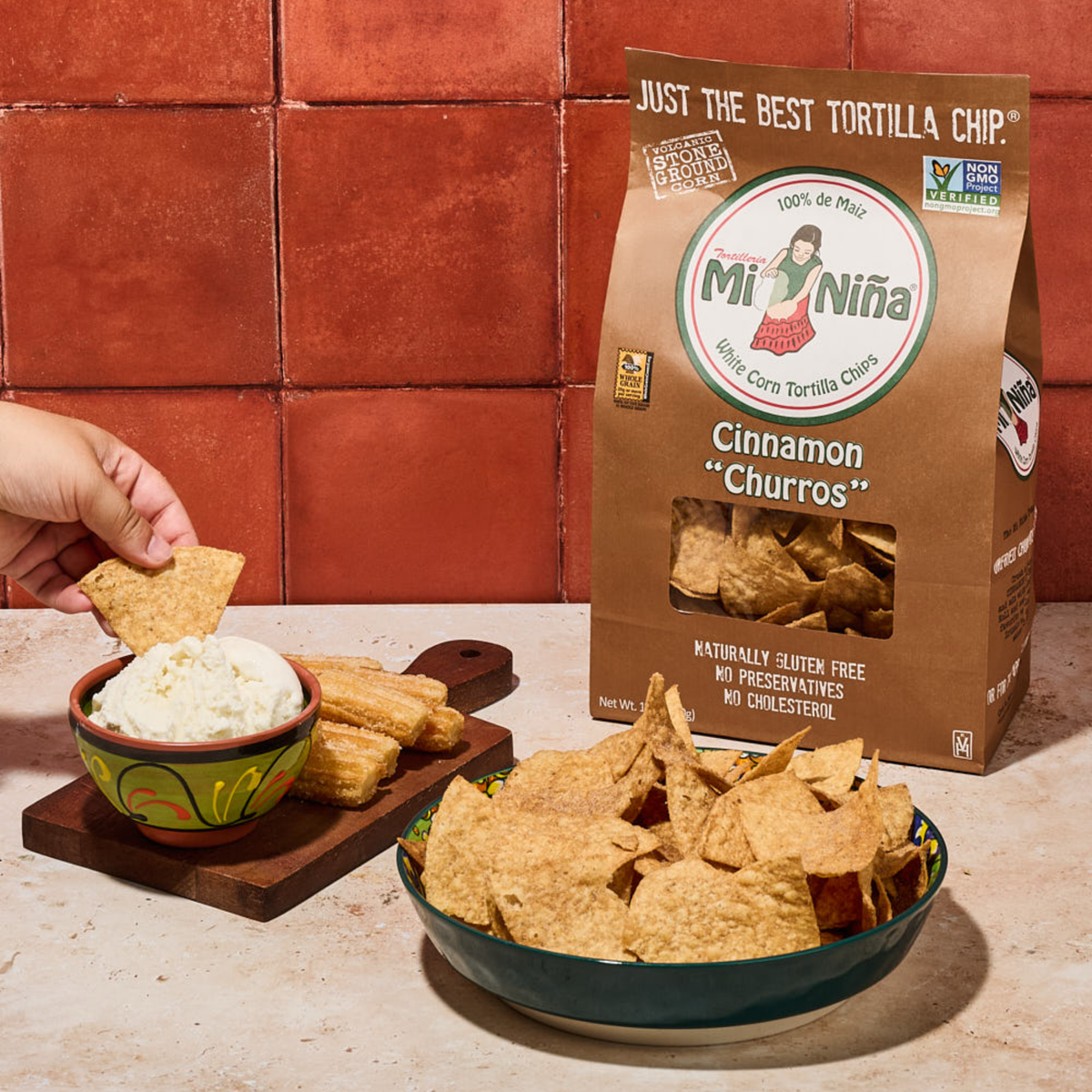 A hand dips a white corn tortilla chip into a bowl of white dip. On the counter against a red tile wall, there is a large bowl of chips, some churros, and a 12 oz bag of Mi Niña Tortilla Cinnamon “Churros” Tortilla Chips (9 Pack).