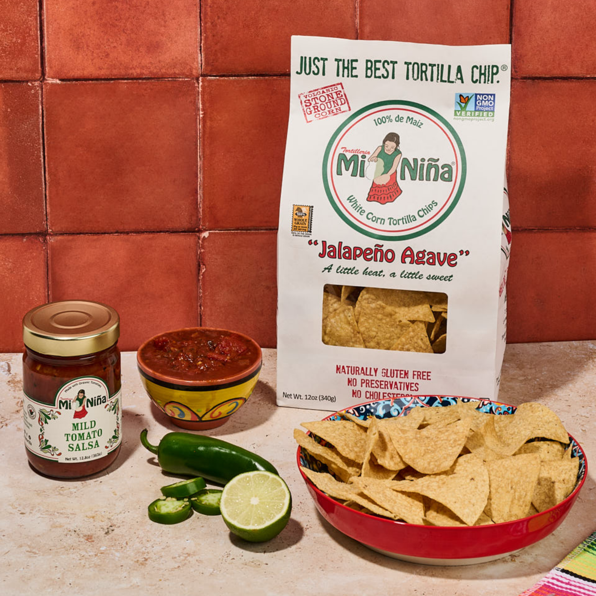 A bag of Mi Niña Tortilla “Jalapeño Agave” Tortilla Chips (12 oz), a bowl filled with the chips, a jar of mild tomato salsa, a small bowl of salsa, two fresh jalapeños, and half a lime arranged on a tiled counter.