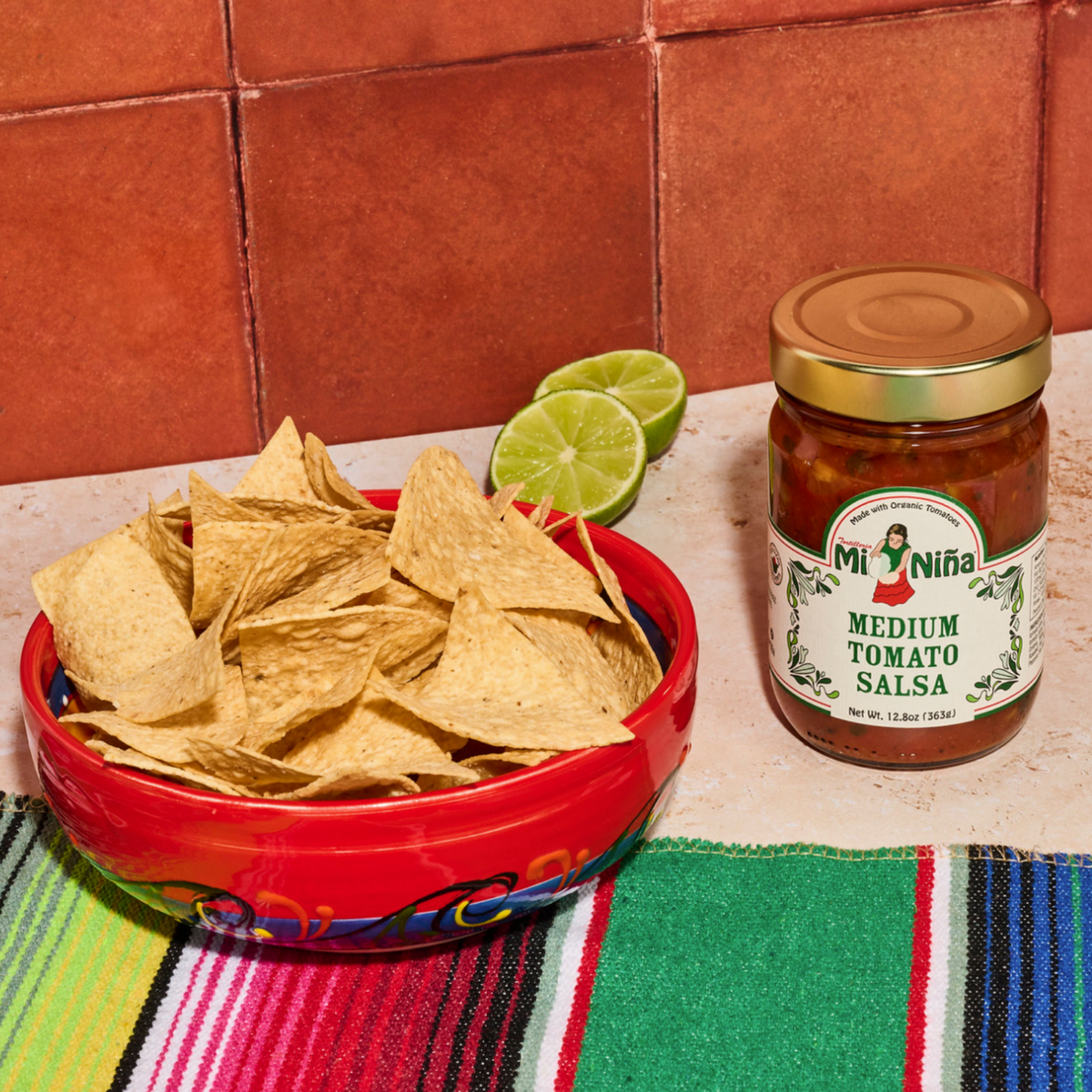 A bowl of tortilla chips sits next to a jar of Mi Niña Tortilla Medium Tomato Salsa (12.8 oz, from the 6 Pack). A cut lime and a colorful striped cloth are nearby, all set on a tiled surface.