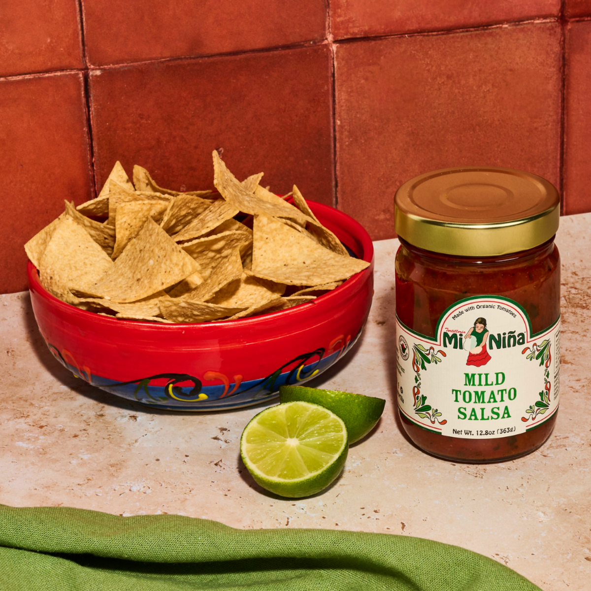 A vibrant bowl of tortilla chips sits beside a sliced lime and a 12.8 oz jar of Mi Niña Tortilla Mild Tomato Salsa on the countertop, with a green cloth and terracotta tiles in the background. Perfect for parties, this Mild Tomato Salsa is also available in a convenient 6 pack.