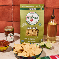 A bowl of white corn tortilla chips, a bag of Mi Niña Tortilla Sea Salt Made with Olive Oil Tortilla Chips (9 Pack / 10 oz), a jar of salsa, an olive oil bottle, lime halves, and a colorful napkin are arranged on a table with a red tile background.