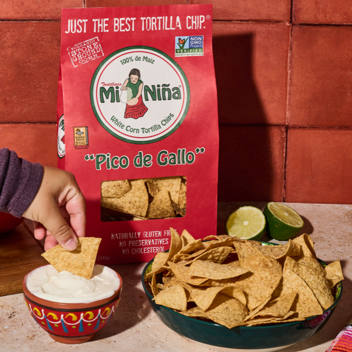 A hand dips a Mi Niña Tortilla “Pico de Gallo” Tortilla Chip into sour cream, next to a bowl of tortilla chips, a halved lime, and a red bag labeled Mi Niña “Pico de Gallo” Tortilla Chip - 9 Pack / 12 oz.