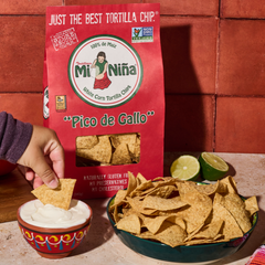 A hand dips a white corn tortilla chip into a small bowl of pico de gallo next to a large bag of Mi Niña Tortilla Pico de Gallo Tortilla Chips – 12 oz Bags (9-Pack) and lime halves.