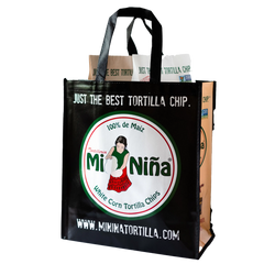 A black Reusable Tote Bag from Mi Niña Tortilla features a logo of a woman holding chips and is filled with brown paper tortilla chip bags. The text reads “Just the Best Tortilla Chip” along with the website www.mininatortilla.com.