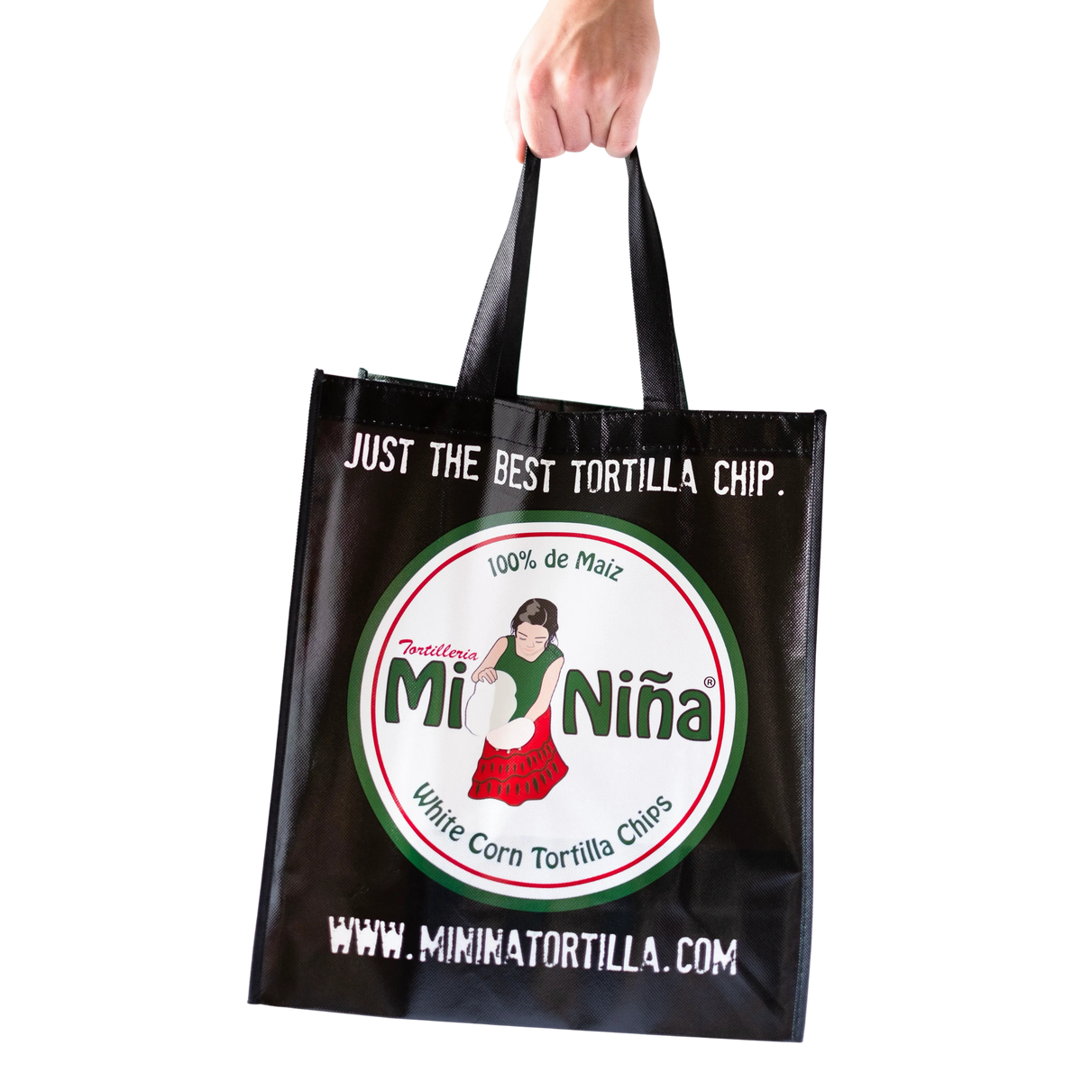 A hand holds the Mi Niña Tortilla Reusable Tote Bag in black, featuring the Mi Niña logo with a woman holding corn tortillas and the phrases “Just the best tortilla chip” and “www.mininiatortilla.com” printed on it.