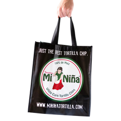 A hand holds the Mi Niña Tortilla Reusable Tote Bag in black, featuring the Mi Niña logo with a woman holding corn tortillas and the phrases “Just the best tortilla chip” and “www.mininiatortilla.com” printed on it.