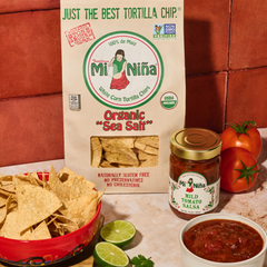 A bag of Mi Niño Tortilla Organic “Sea Salt” White Corn Tortilla Chips (12 oz), an open jar of mild tomato salsa, a bowl filled with chips, fresh tomatoes, and lime slices sit on a kitchen counter with a red tile backsplash.