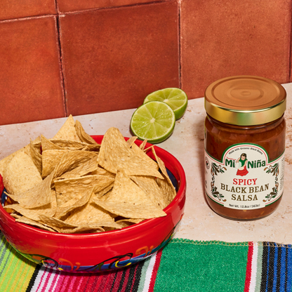 A bowl of tortilla chips sits next to a 12.8 oz jar of Mi Niña Tortilla Spicy Black Bean Salsa – 6 Pack, accompanied by sliced limes and a colorful striped cloth on a tiled surface.