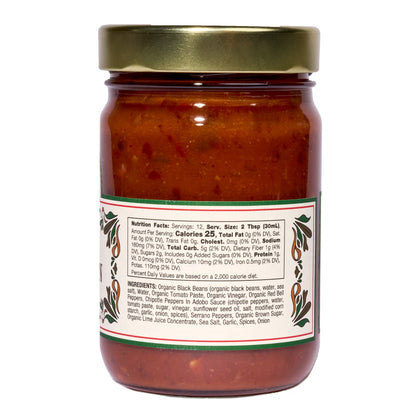 A clear 12.8 oz glass jar of Mi Niña Tortilla Spicy Black Bean Salsa, sealed with a gold lid. The back label shows nutrition facts and ingredients in black on white with green and red accents. Sold in a convenient 6 pack.