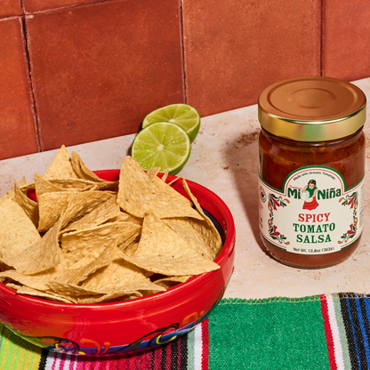 A bowl of tortilla chips sits beside a jar of Mi Niño Tortilla Spicy Tomato Salsa (12.8 oz)—available in a convenient 6 pack—accompanied by two lime halves and a colorful striped cloth on a tiled surface, making it ideal for sharing or stocking up.