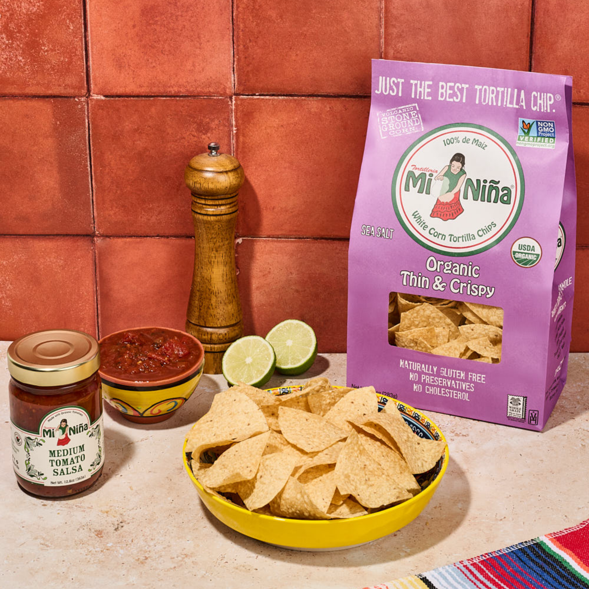 A yellow bowl filled with thin & crispy tortilla chips, a jar of salsa, a small bowl of salsa, lime halves, and a pepper mill are arranged on a tiled countertop with a purple Mi Niña Tortilla Organic Thin & Crispy Sea Salt Tortilla Chip bag (9 Pack / 10 oz) in the background.