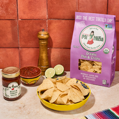 A yellow bowl filled with thin & crispy tortilla chips, a jar of salsa, a small bowl of salsa, lime halves, and a pepper mill are arranged on a tiled countertop with a purple Mi Niña Tortilla Organic Thin & Crispy Sea Salt Tortilla Chip bag (9 Pack / 10 oz) in the background.