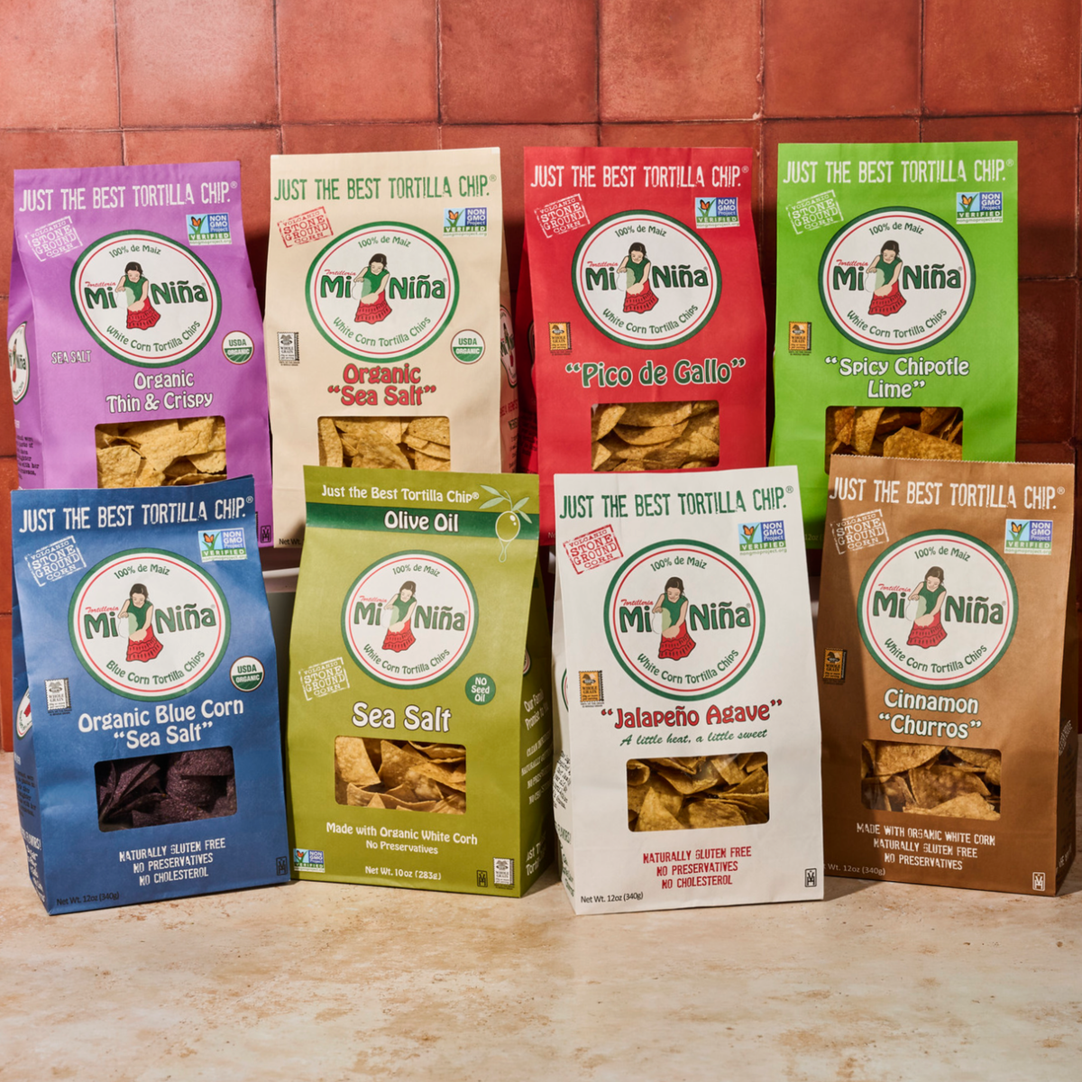Three bags of Mi Niña Tortilla Variety Pack (3 bags) featuring assorted flavors like sea salt, pico de gallo, and spicy chipotle lime are displayed on a tiled surface with a red-brown background.